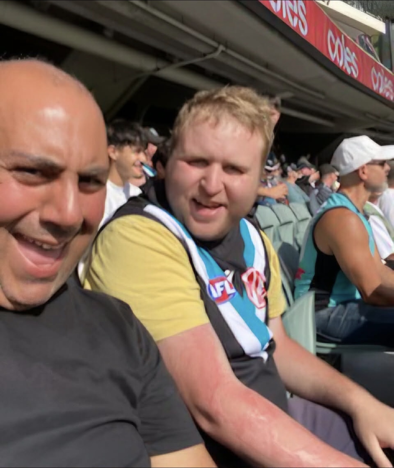 Two mates in the crowd at the footy on a sunny afternoon, one in his team guernsey, both smiling